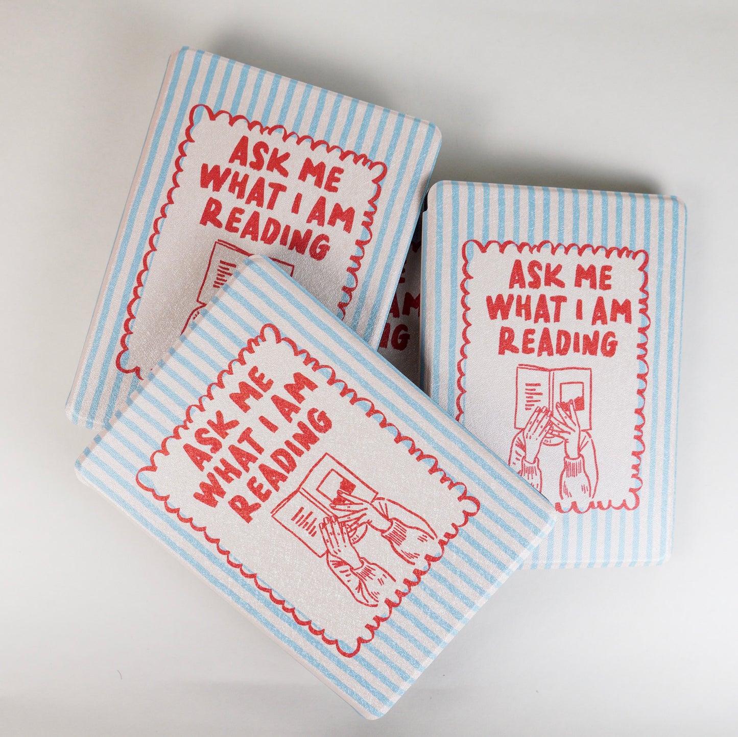 A pile of Page Turner Kindle Case with blue and beige stripes, red text that says ‘Ask Me What I’m Reading,’ and hands holding a book illustration by Elco House