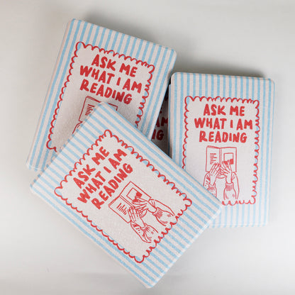 A pile of Page Turner Kindle Case with blue and beige stripes, red text that says ‘Ask Me What I’m Reading,’ and hands holding a book illustration by Elco House